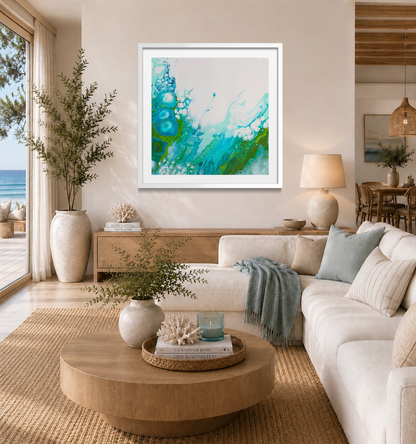 A framed abstract painting with vibrant teal, green, and blue brushstrokes on a white background, displayed on a beige wall in a modern, sunlit living room with a white sofa and wooden coffee table.