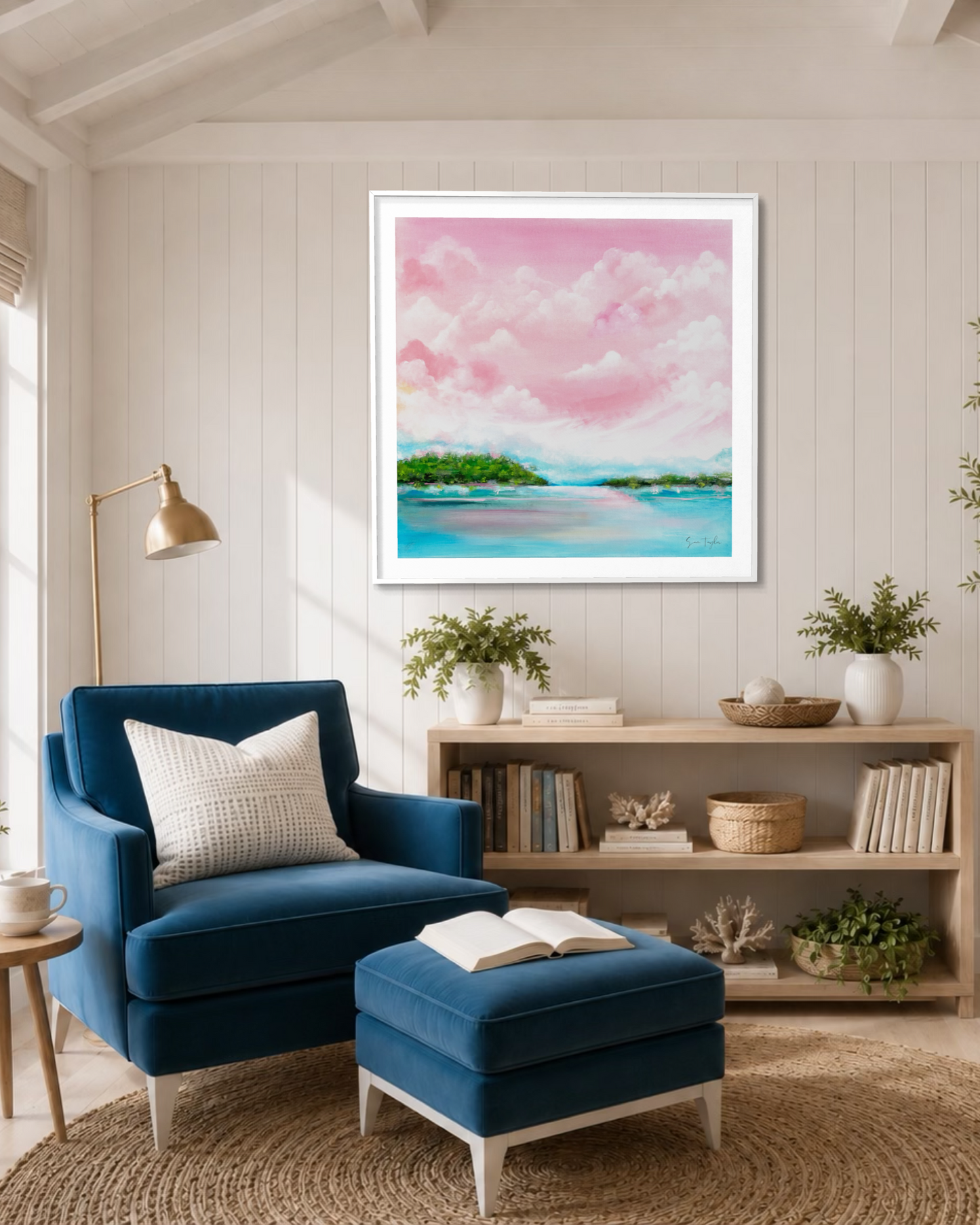 A framed square art print depicting a pink and blue tropical beach landscape hangs on a white paneled wall above a blue armchair and matching ottoman in a bright, modern living room.