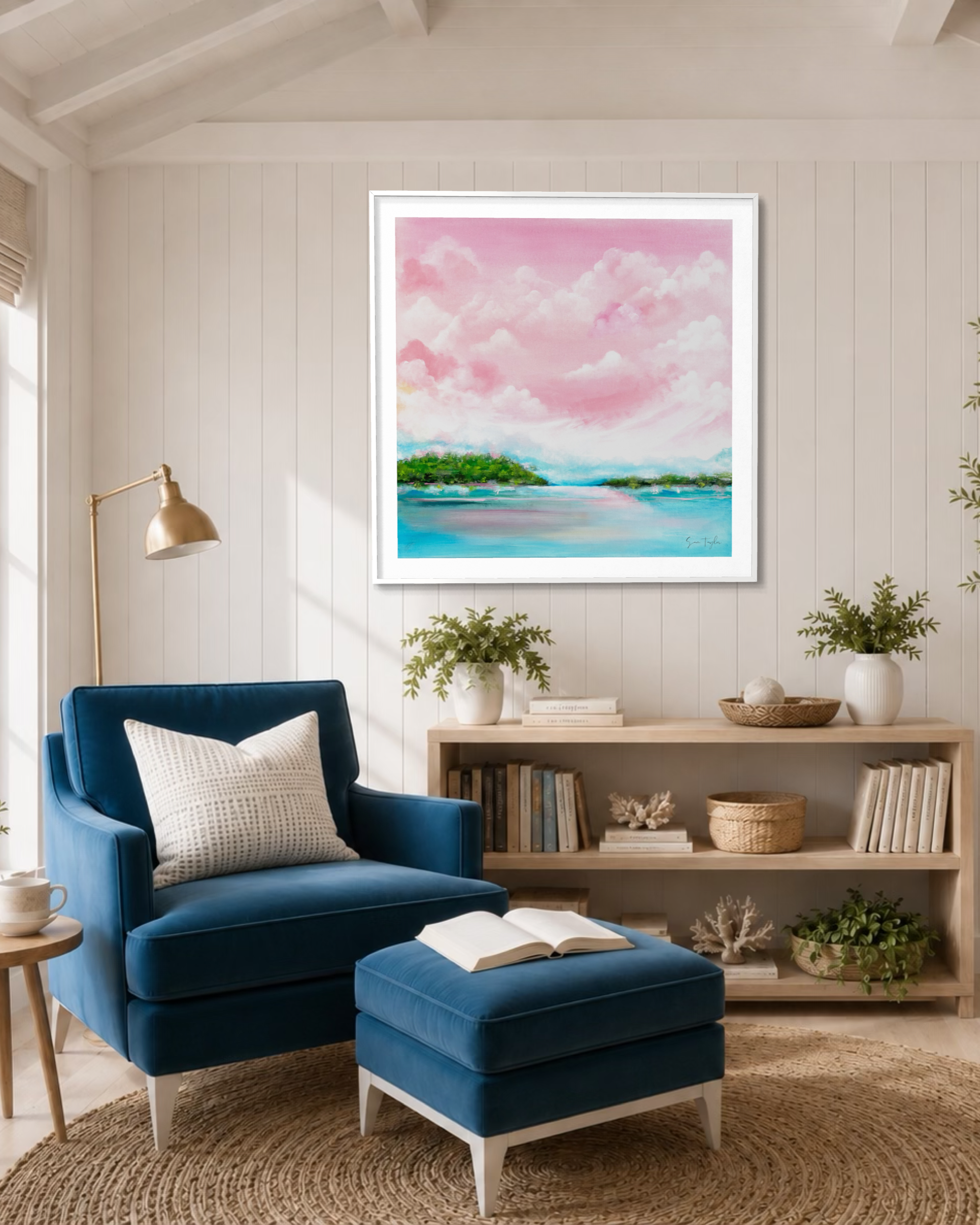 A framed square art print depicting a pink and blue tropical beach landscape hangs on a white paneled wall above a blue armchair and matching ottoman in a bright, modern living room.