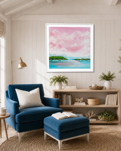 A framed square art print depicting a pink and blue tropical beach landscape hangs on a white paneled wall above a blue armchair and matching ottoman in a bright, modern living room.