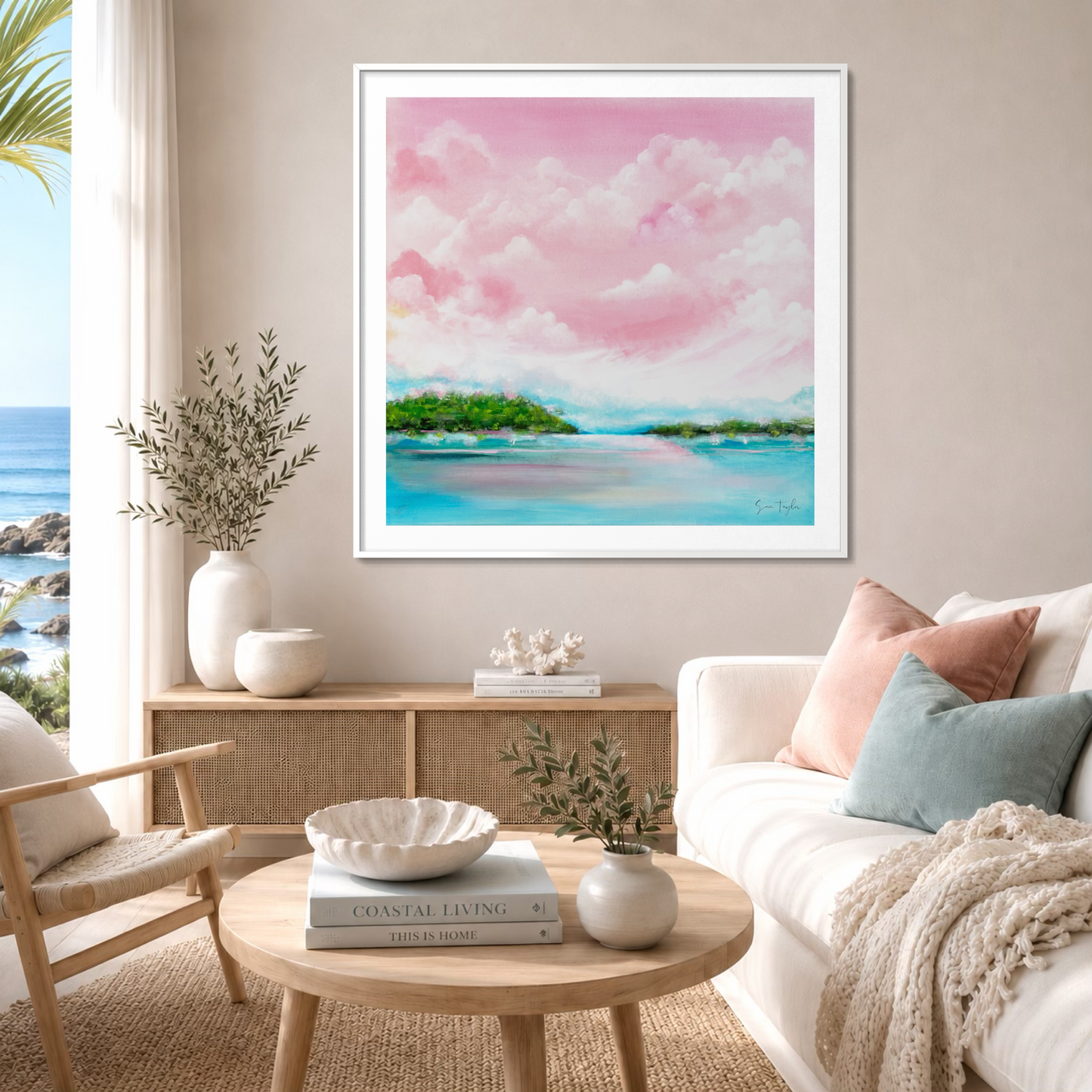 A framed pastel landscape painting of a tropical beach with pink clouds, turquoise water, and green islands, displayed on a beige wall above a wooden coffee table in a bright, modern living room.