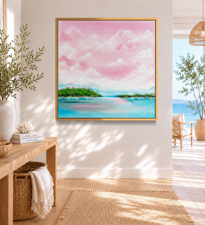 A framed square canvas painting of a pink sky with white clouds, turquoise water, and green islands, displayed on a white wall in a bright, modern living room interior.