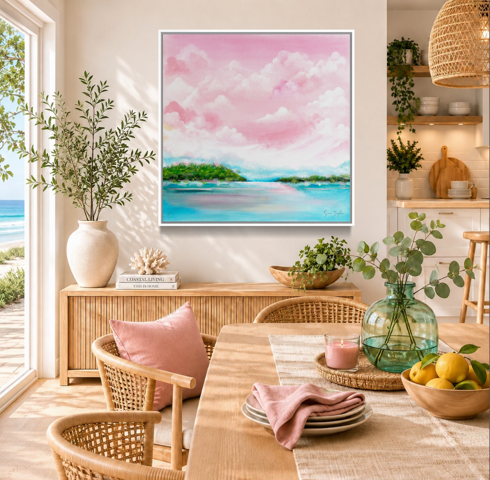 A framed pastel landscape painting of pink clouds and turquoise water is displayed on a wall in a bright, modern dining room with wicker chairs, a wooden table, and a vase of greenery.