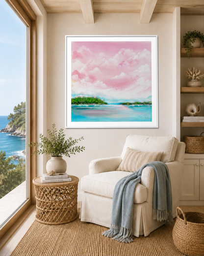 A framed pastel landscape painting of pink clouds and turquoise water with green islands, displayed on a beige wall above a cream armchair in a bright coastal-style room.