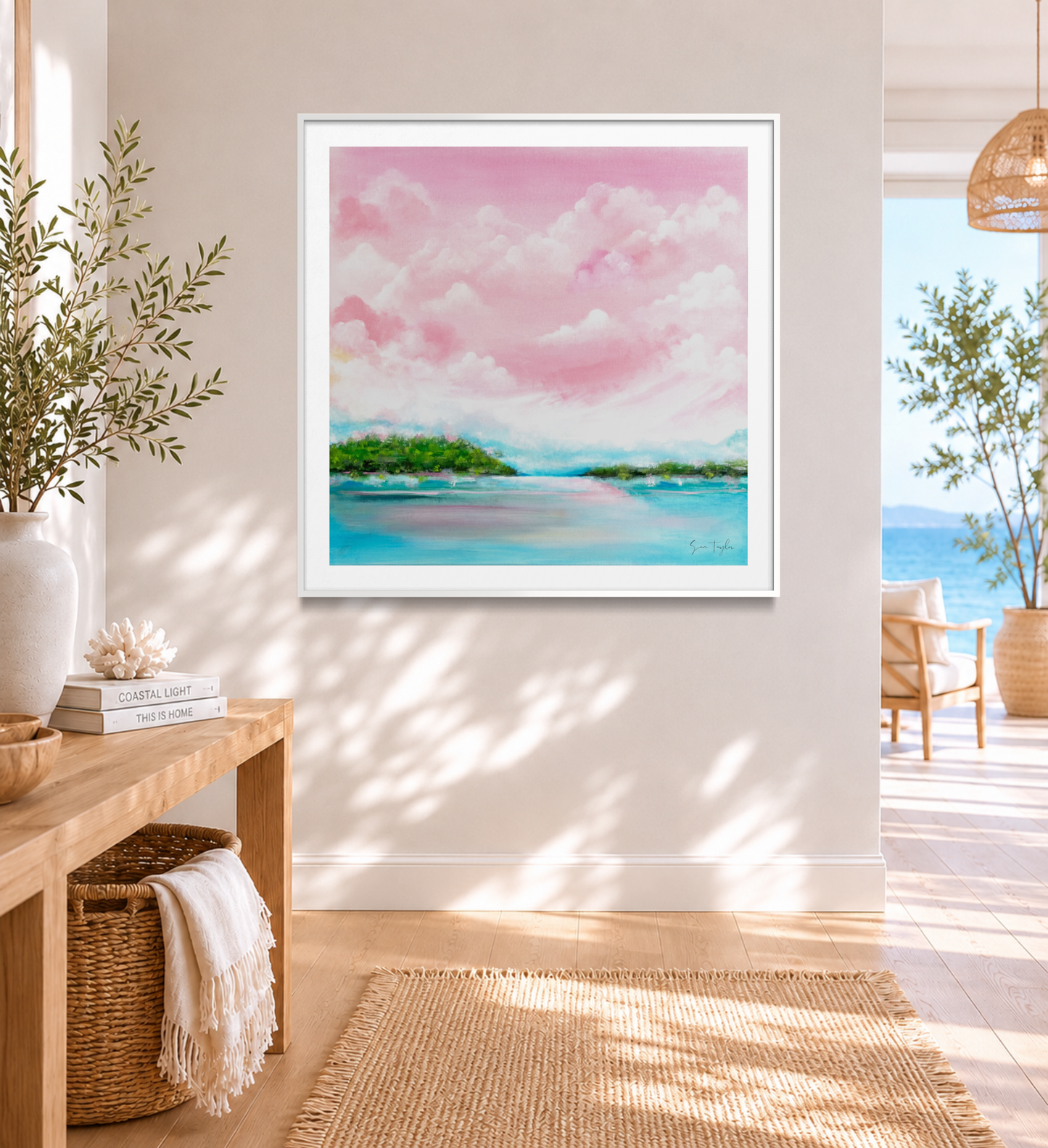 A framed square art print depicting a pastel pink sky over turquoise water and green islands, displayed on a light beige wall in a bright, modern living room with a wooden console table and woven rug.